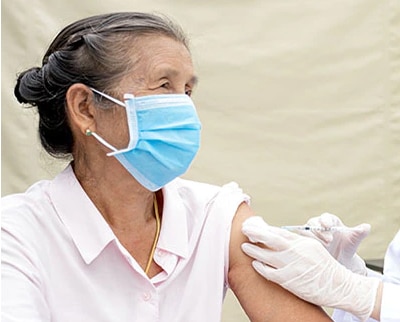 An older female patient wearing a masks sits while a health care provider administers a vaccine in her arm.