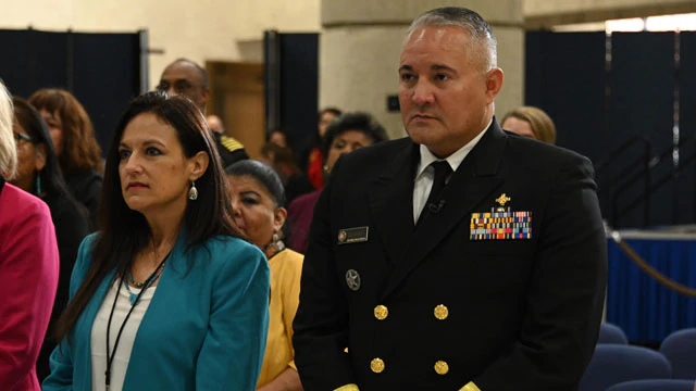 A person in a highly decorated military uniform stands next to others in an audience