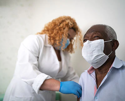 An older male patient wearing a masks sits while a female health care provider wearing a mask and gloves administers a vaccine in his arm.