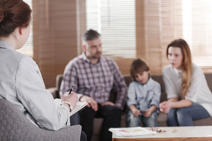 A case worker takes notes while talking to two parents and their child, who are sitting on a couch in the background.