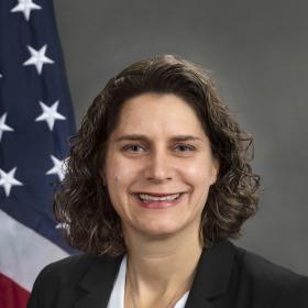 Alison Barkoff is seated in a dark suite, white blouse with American flag behind her.