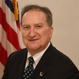 Bob Valdez is seated in front of a American flag wearing a dark colored suite.