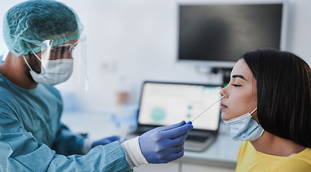 Medical provider wearing personal protective equipment performs coronavirus nose swab on young adult patient.