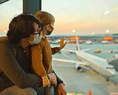 Adult with child wearing masks looking out window at an airplane in an airport.