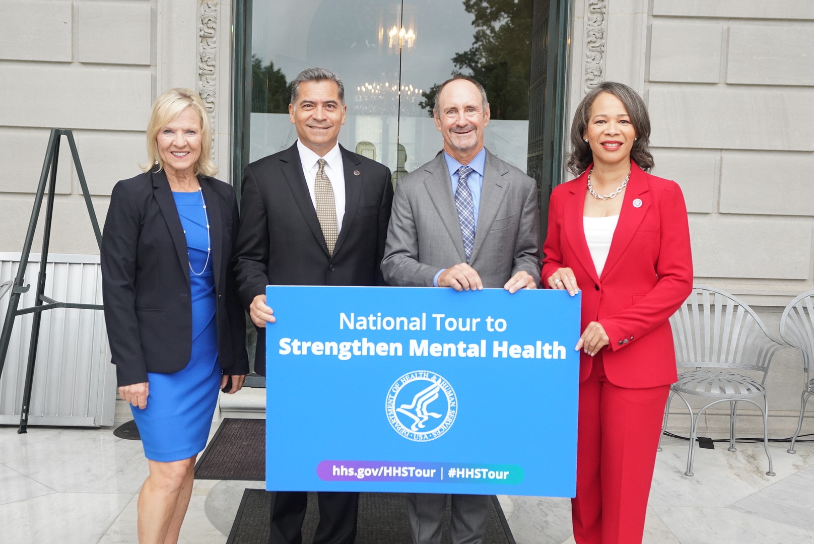 Secretary Becerra is standing behind a light blue Mental Health tour sign wearing a dark suit. To the right of him is a male wearing a light grey suit with a blue shirt, to the right of him is a lady wearing a red pant suit. To the left of Secretary Becerra is a lady wearing a blue dress with a black blazer.